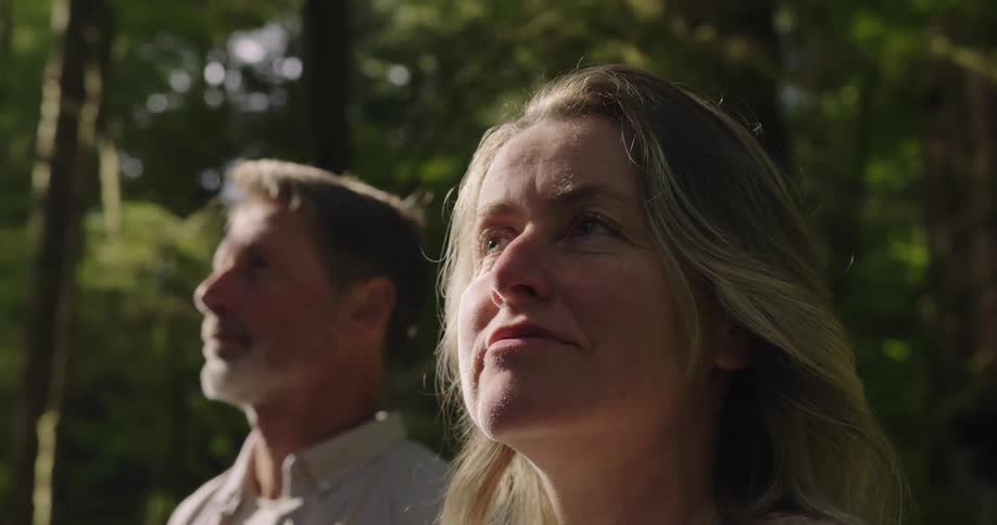 A woman and a man stand sidebyside in a forest both looking up and to the side