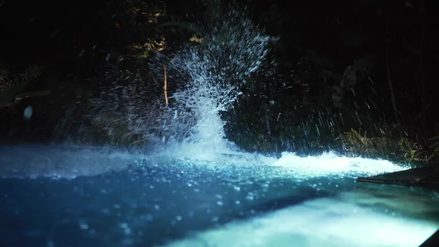 The video shows a man and a woman in white clothes holding hands as they jump into a swimming pool at night they land simultaneously creating a large dramatic splash and then surface in the glowing blue water the scene is lit by the pools lights cr