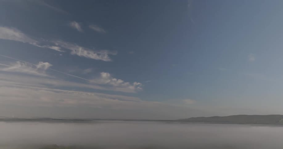 A full view aerial shot shows a thick blanket of white fog blanketing a valley with small hills visible in the distance under a pale blue sky