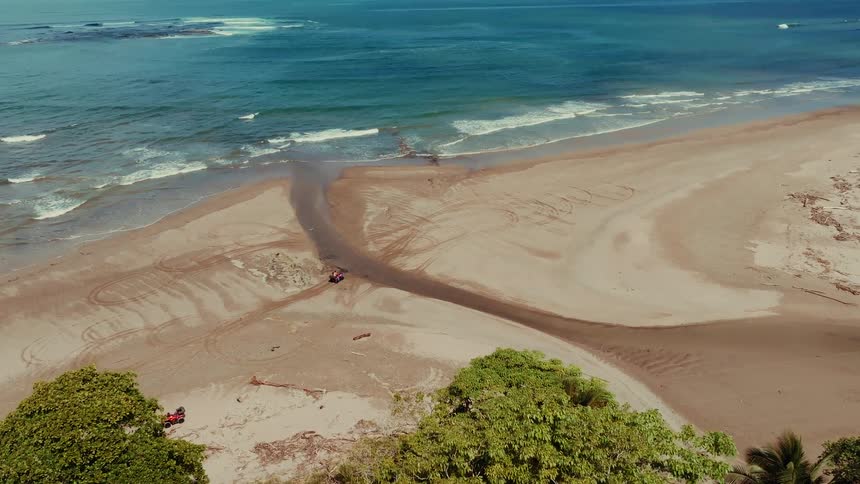 An aerial view shows waves meeting a wide sandy beach where a narrow stream winds through toward the ocean