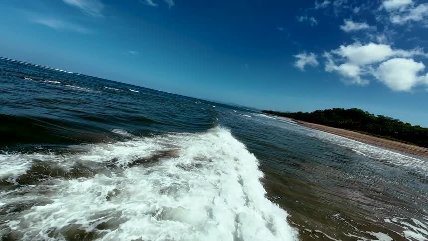 An aerial shot flies over the water as a wave rolls toward a sandy beach