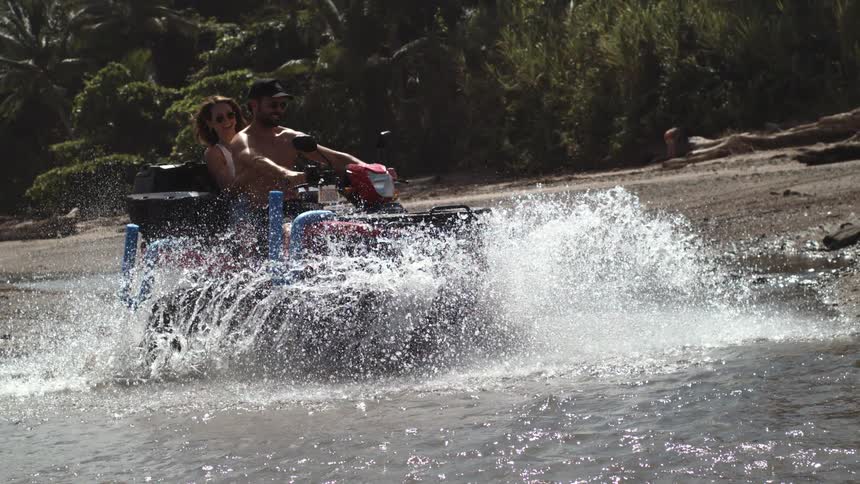 Couple riding a red atv through shallow beach water at midday with the shirtless young man wearing a hat and shorts as the woman rides behind him