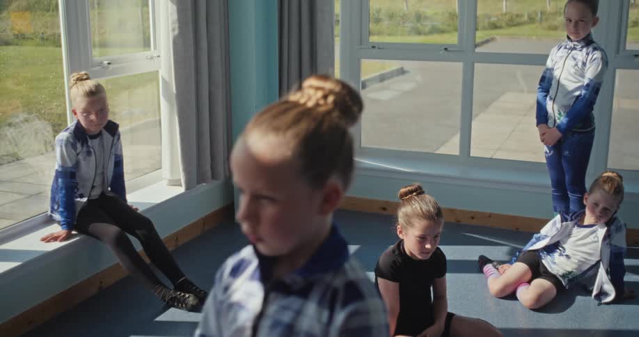 The video shows four young children in a dance or gymnastics studio they are all sitting or leaning in a relaxed pose with their hair tied in a bun waiting for the next instruction