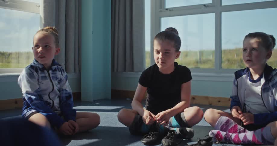 A group of children sits on a floor and talks with each other while an adult comes behind them in an indoor setting