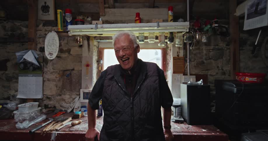 Smiling elderly man in a rustic workshop surrounded by tools and work items
