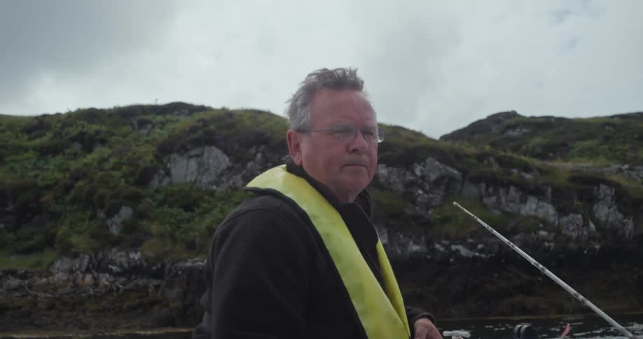 In this short clip a man with glasses and a graying beard is wearing a life vest he is on a boat looking toward the left side of the frame with a rocky green hillside in the background