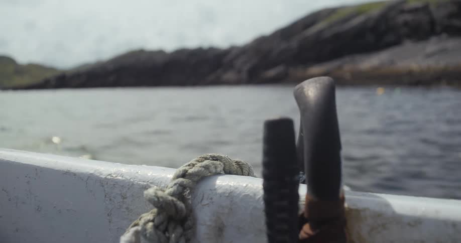 A boat is shown from the bow where a thick rope is tied to the side in the background there is a large body of water surrounded by grassy rocky hills under a bright overcast sky