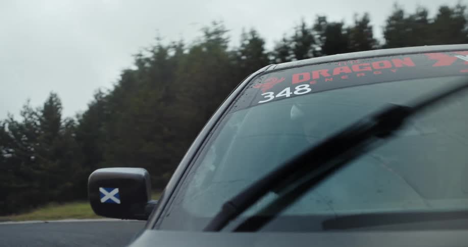 A rally car with the number 348 and a scottish flag on its mirror drives along a race track under an overcast sky