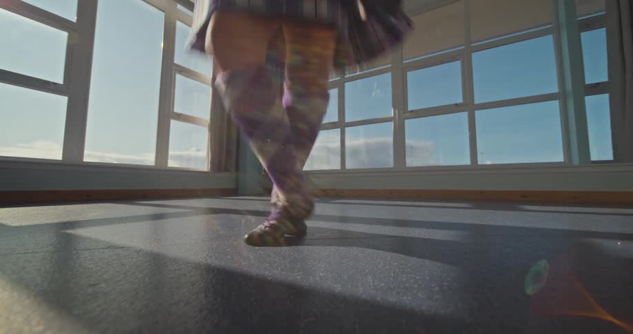 A dancer wearing a short plaid skirt and long patterned socks is seen from the knees down performing a traditional scottish dance the dancers feet are moving quickly and their skirt and socks are visible as they move across a dark floor
