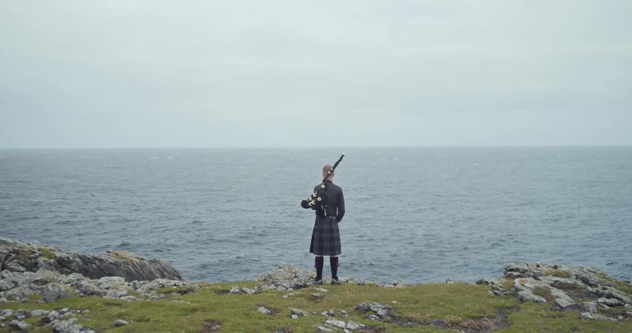 A young boy stands on a grassy cliffside playing a bagpipe while facing the ocean