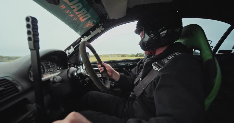 A test driver with full protective gear including a helmet and reinforced safety belt at the wheel of a car