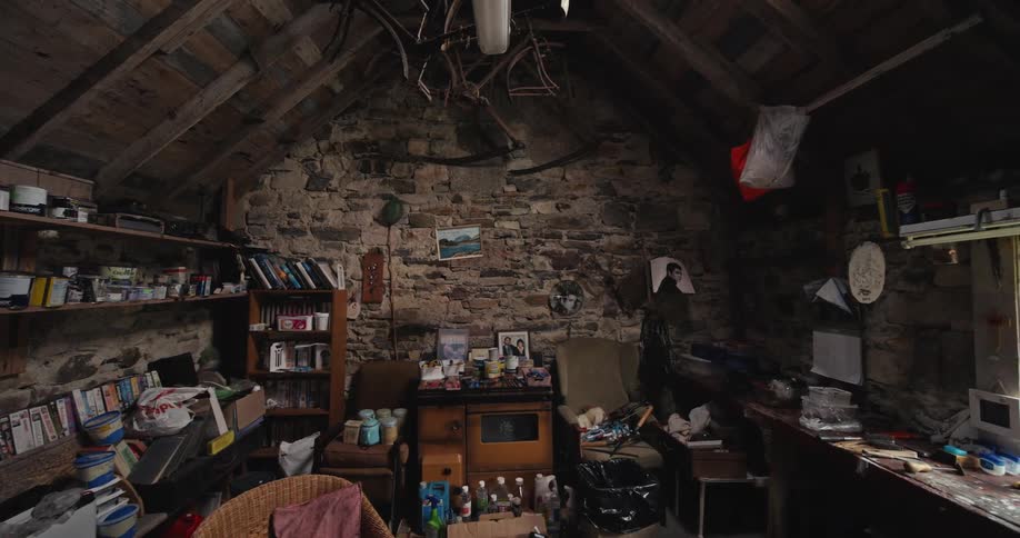 A slow panning wide view of a cluttered workshop with stone walls wooden rafters and an assortment of tools shelves and furniture