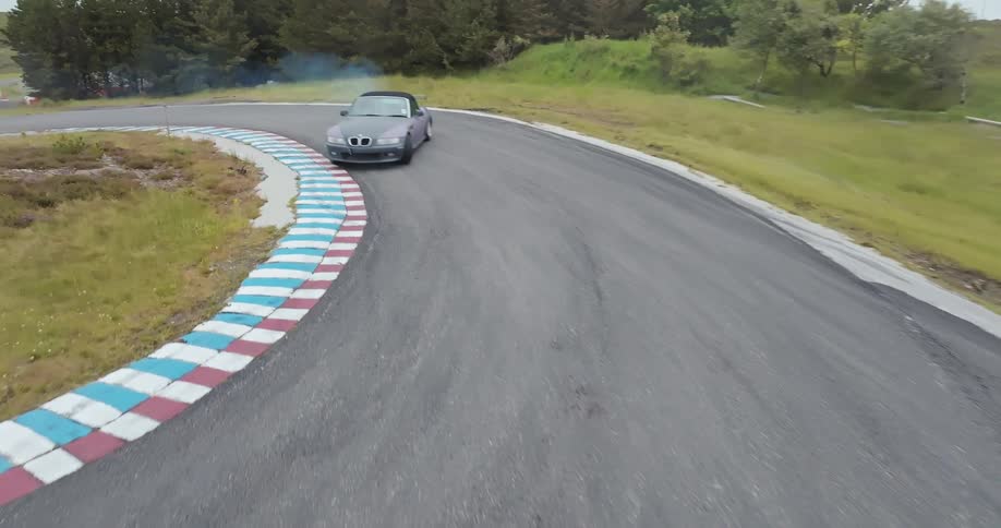 A dark gray sports car is seen driving on a race track drifting and creating smoke from its tires as it navigates a turn the car is on a track with a red white and blue curb on the inside of the curve