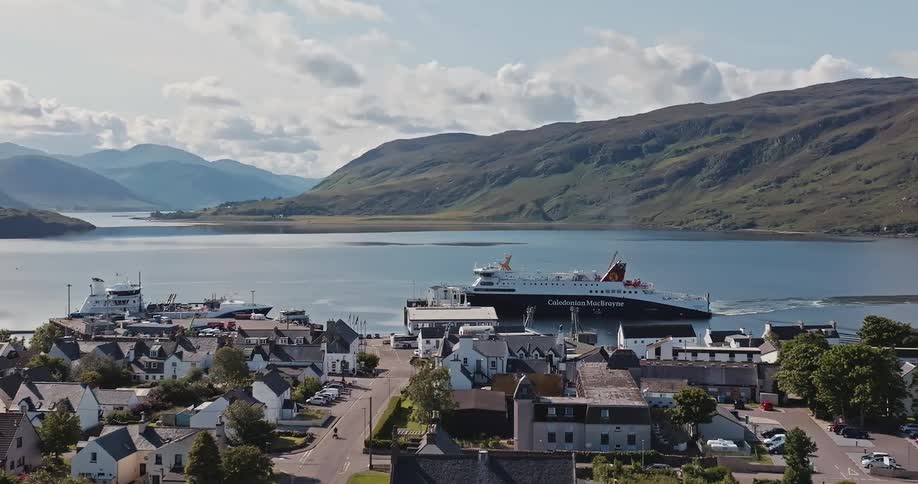 A large ferry approaches a quaint port town nestled against a mountainous bay under a cloudy sky
