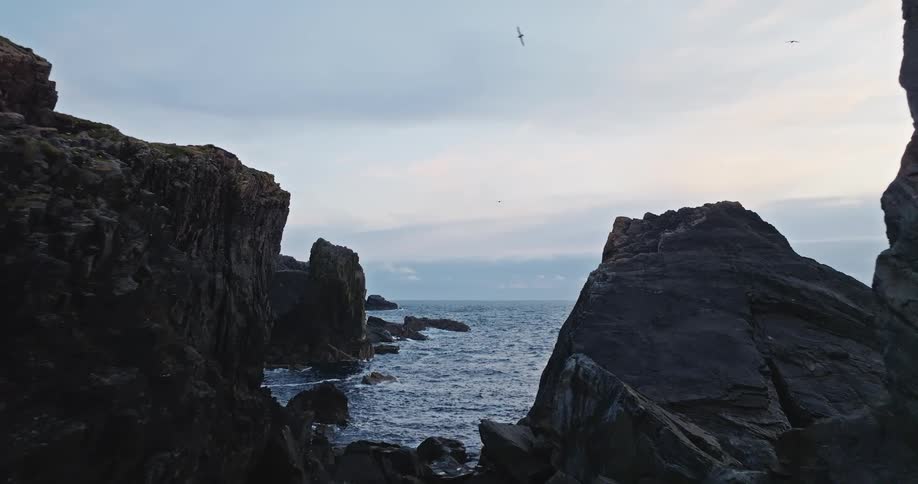 The scene is an ocean inlet with waves gently crashing against the shore and large rocky cliffs framing the view the sky is overcast and a bird can be seen flying across the horizon adding a sense of movement to the otherwise calm scenery