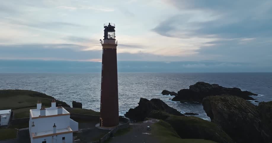 The video shows an aerial shot of a lighthouse situated on a rocky coastline the camera slowly zooms in on the lighthouse which stands tall and is surrounded by a few small white buildings the waves of the dark blue ocean crash against the rocks below
