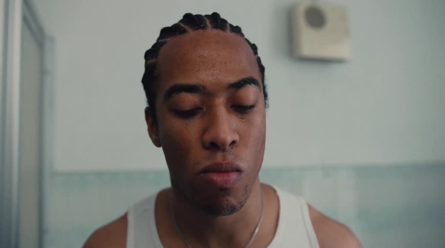 A man with braided hair and a tank top looks directly at the camera then moves his hands over his face appearing contemplative or tired