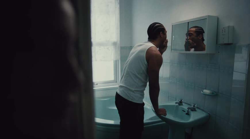 A man wearing a white tank top and dark pants is washing his face in a bathroom sink after splashing water on his face he looks up into the mirror staring at his reflection