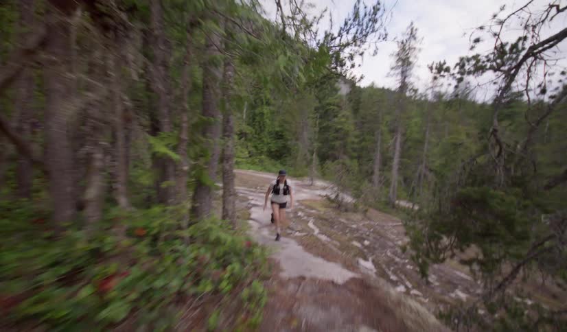 A low angle view from behind a woman in a tank top and black shorts shows her running up a narrow rocky trail in a dense green forest