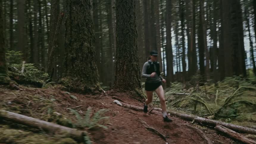 A full view of a woman wearing a black tank top and shorts running down a steep dirt path through a forest of tall dense trees