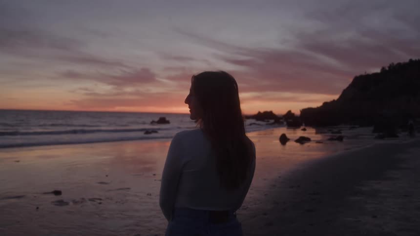 A person with long hair stands on a beach facing away from the camera with the ocean and a cliff behind them as the sun sets