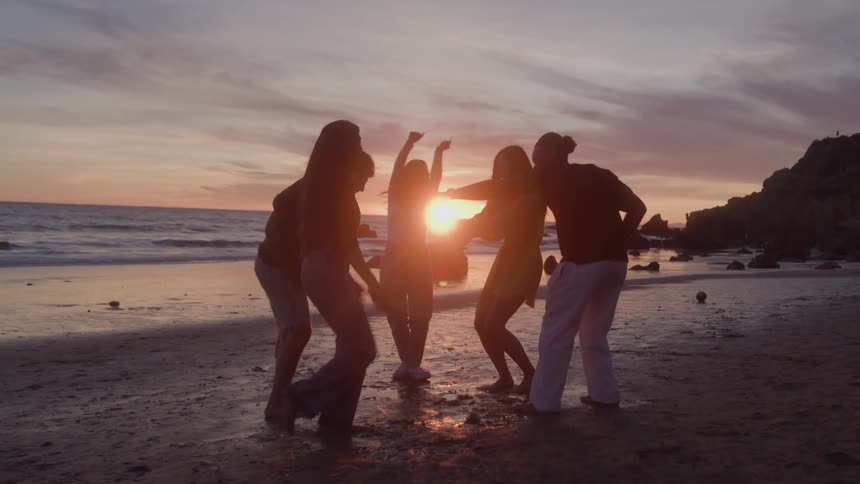 A group of people stand on a beach facing each other in a loose circle with the ocean behind them as the sun sets
