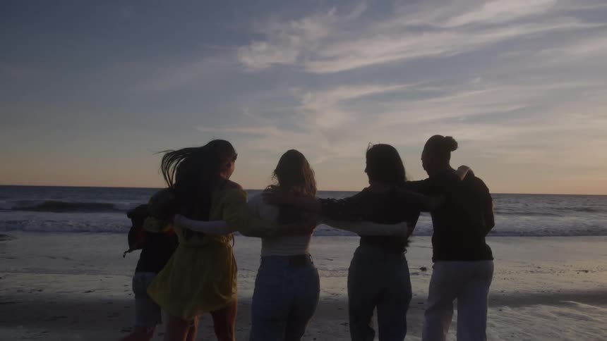 Five friends with arms around each other watching the sea at sunset