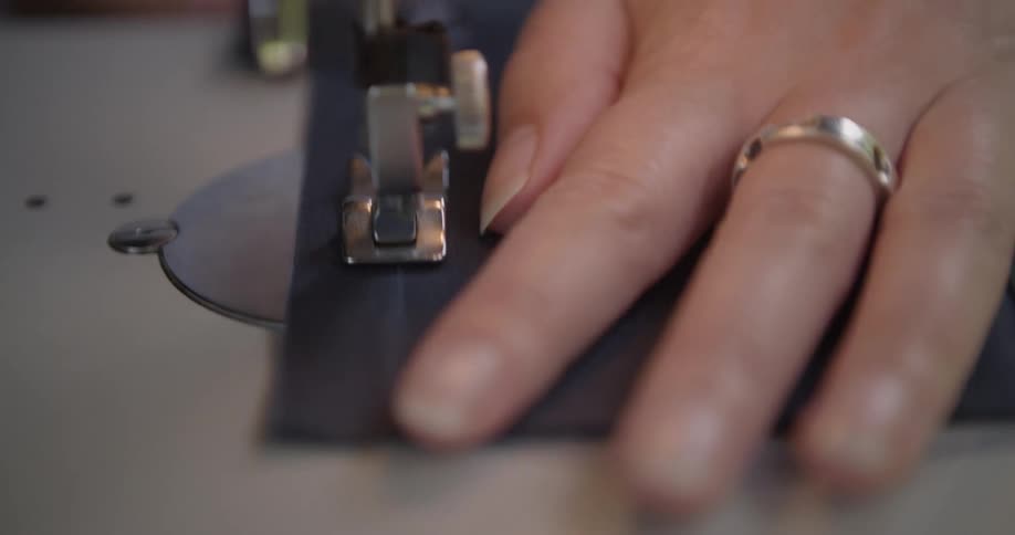 A close view of a persons hands pushing a dark blue fabric through a sewing machine with a shiny metal ring on one finger
