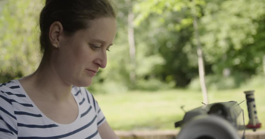 A close view of a woman wearing a striped shirt as she works on a sewing machine outdoors in a bright green yard with lush trees
