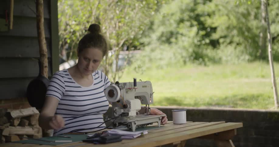 A woman sits at a wood table and uses a sewing machine to sew a piece of material