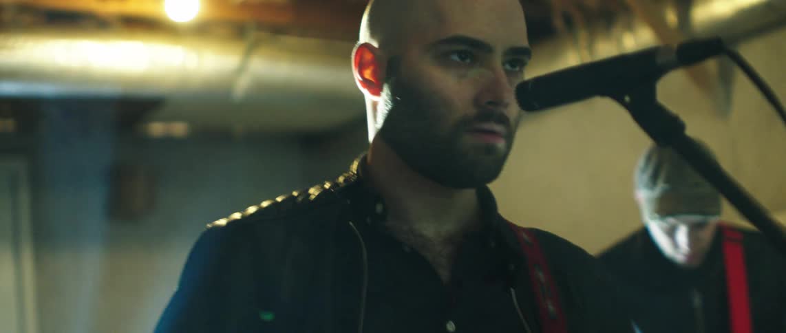 A man singing on a microphone and playing electric guitar with another man playing guitar on the back on a studio setting