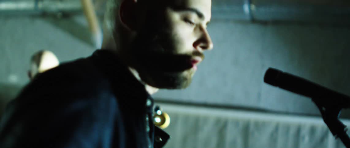 A bald man with a beard passionately screams into a microphone his face lit with intense emotion