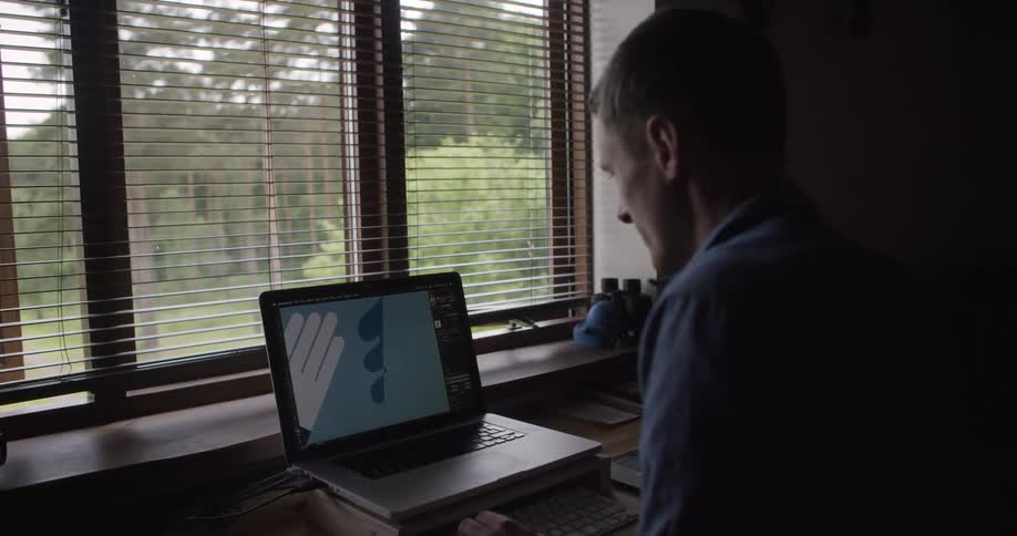 A man works on a laptop with a design program open beside a window