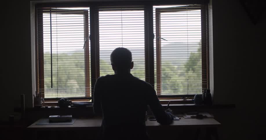 A person is silhouetted as they sit at a desk in front of a large window with open blinds looking out at a hilly landscape