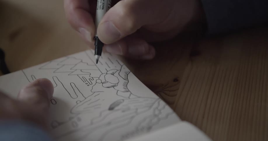 A hand holds a pen and draws lines on a paper in a notebook that is on a wooden table