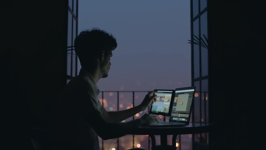 A man is sitting by a window in the dark using a computer