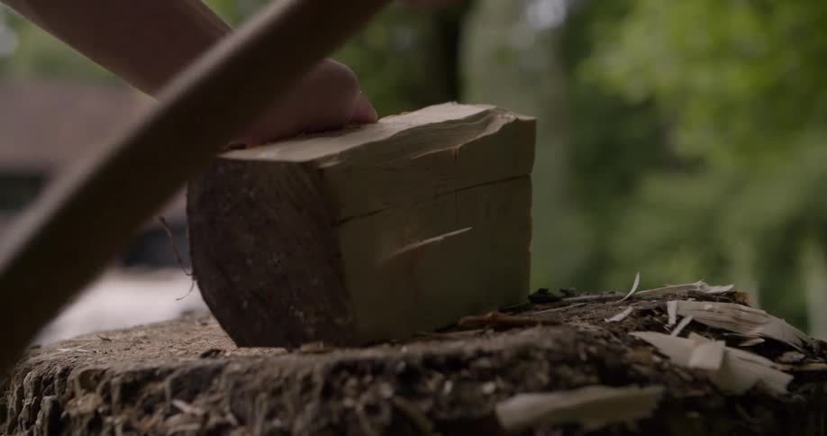 A close view shows a persons hands using a black axe to chop a wooden block on a tree stump in a bright yard