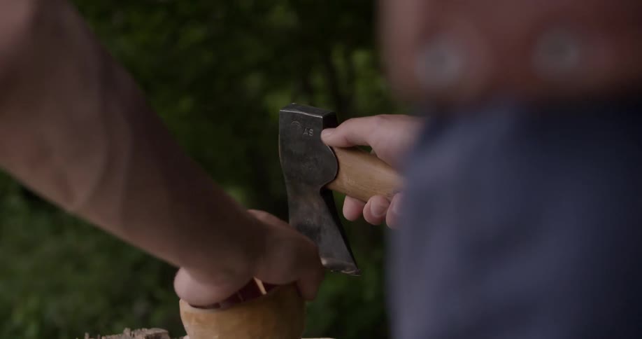 Man uses a hatchet to shape a wooden object on a summer day
