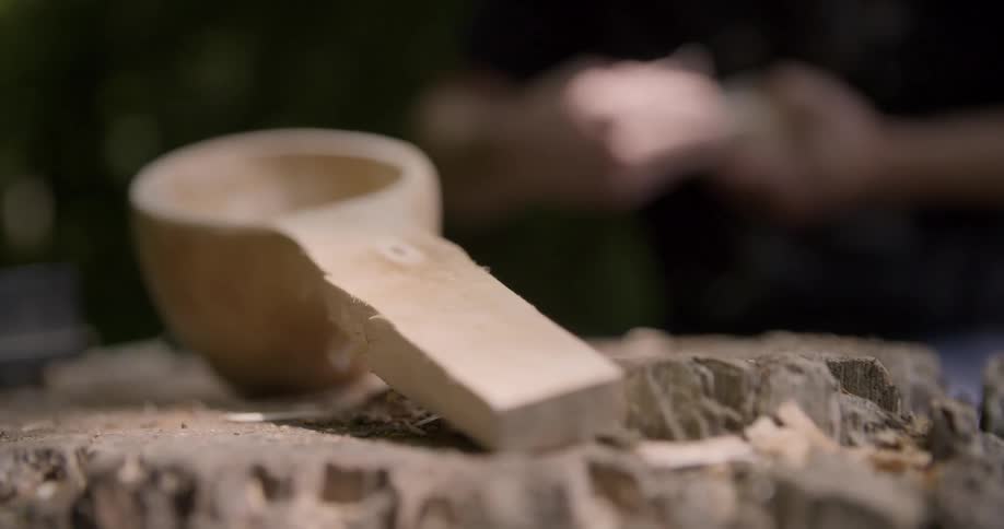 Roughly carved wood scoop lies on an uneven surface while a person smooths another object in the background