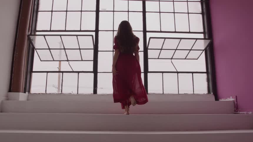 A barefoot woman in a flowing red dress walks up white stairs towards a large window