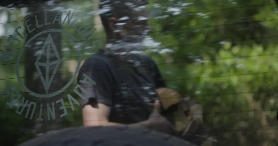 A low angle shot through the rear window of a car shows a man in a black shirt carrying firewood as he walks past