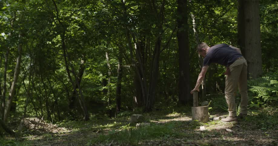 A man in a dark shirt and tan pants chops wood with an axe in a shaded forest clearing