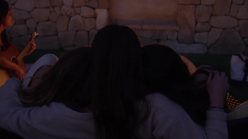 Friends hugging with one of them playing guitar while sitting around a campfire