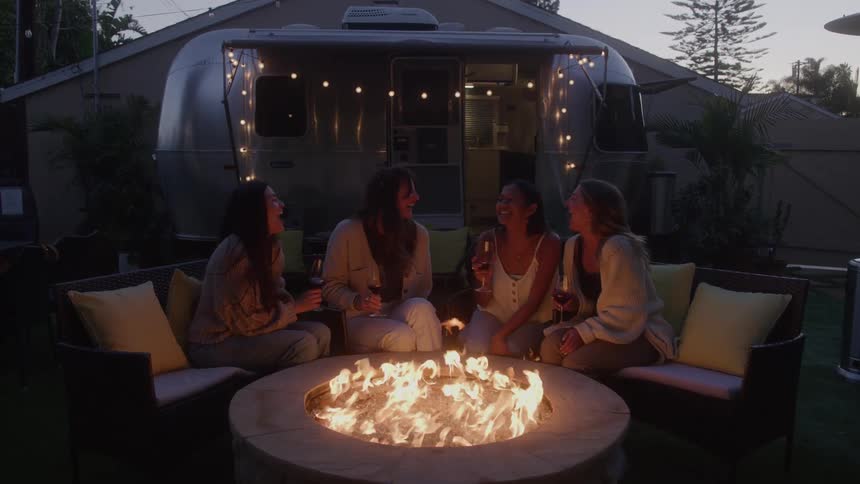 Four friends are gathered around a fire pit on a patio at dusk laughing and enjoying a casual outdoor gettogether the patio is furnished with chairs and in the background a large airstream trailer is visible