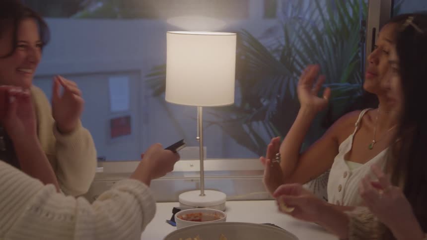 A group of four women are sitting around a table in what appears to be a living room or hotel suite they are talking laughing and eating from a shared bowl of food enjoying their time together a lamp on the table provides a warm soft light