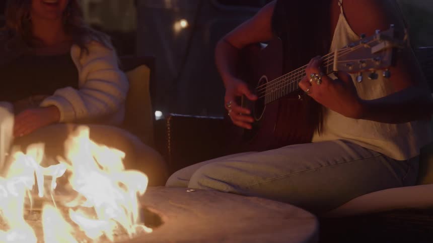 Woman plays guitar while sitting beside the lit fire pit