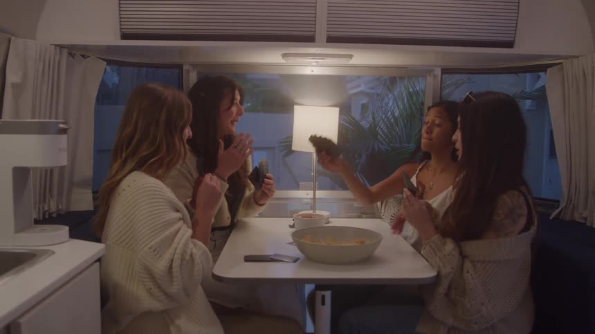 The clip shows a group of four women seated around a table in what appears to be a camper or airstream theyre laughing and looking at their phones then they all raise their hands in the air in an enthusiastic celebratory gesture