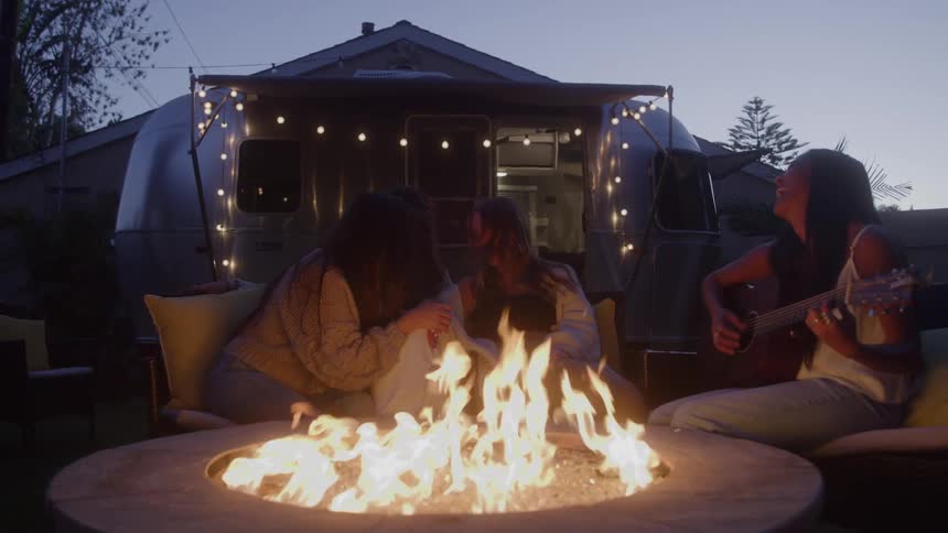 Woman playing guitar and laughing while sitting next to her friends around a campfire