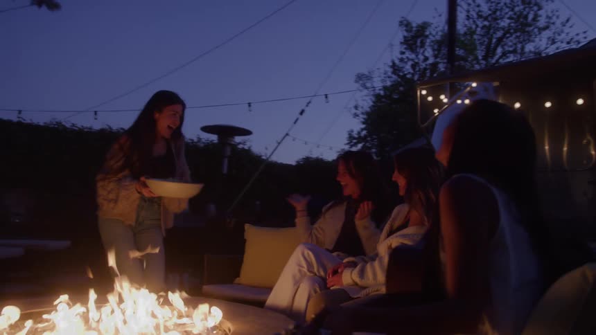 Women gathered around the fire pit smiling while one plays guitar and another arrives bringing food