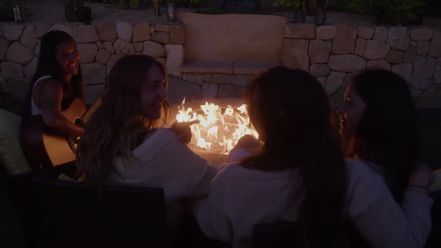 Four women are sitting around a fire pit outdoors with a camper van visible in the background they are all raising their wine glasses smiling and laughing and then taking a sip of their wine as they enjoy each others company